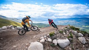 Two mountain bikers ride a corner on the Blue Doon trail at Nevis Range, with an epic mountain view behind them