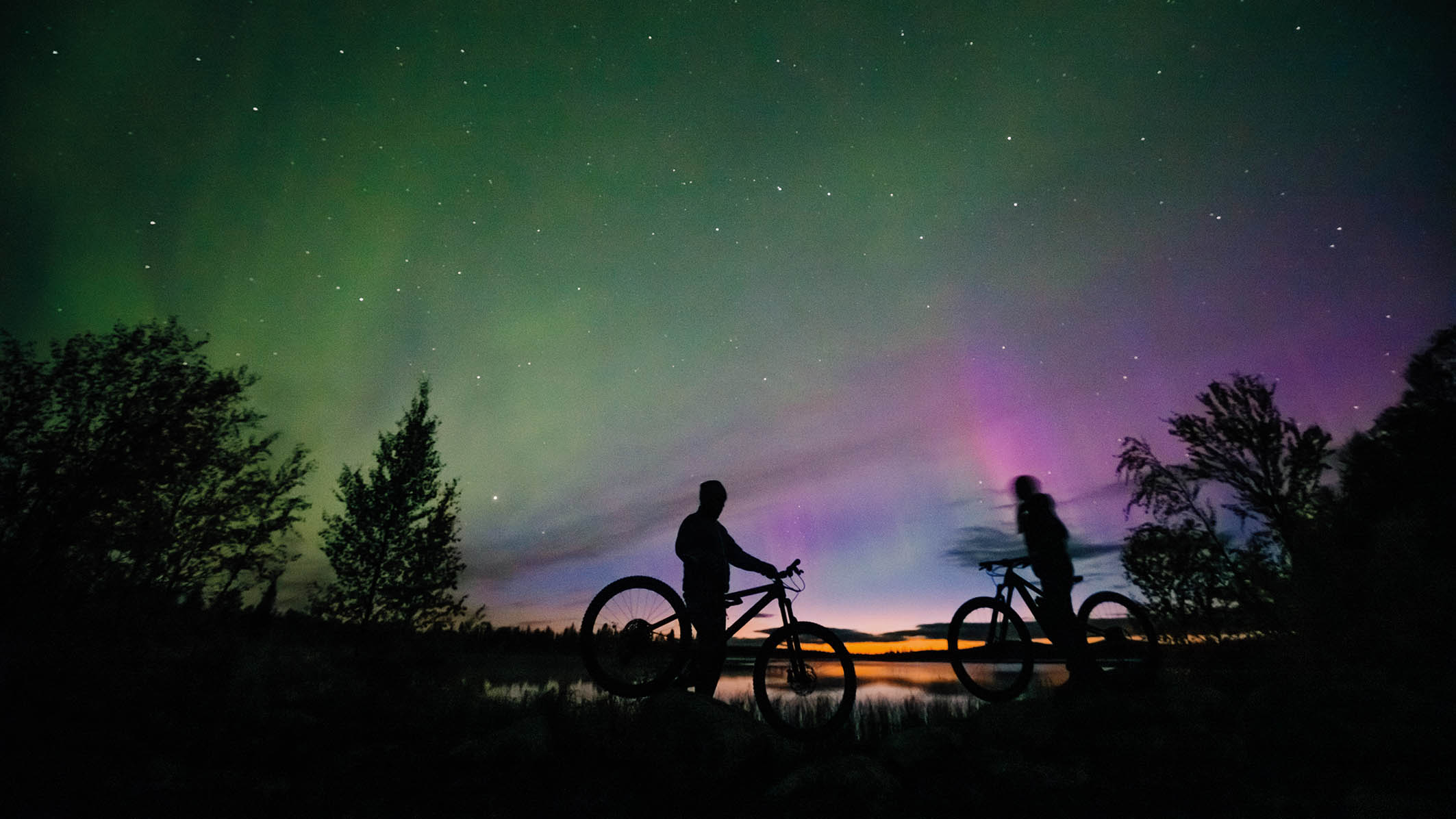 Two mountain bikers in silhouette against northern lights in Lapland