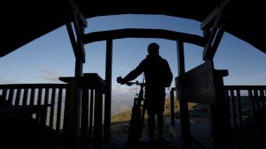 Man silhouetted in wooden structure holding a mountain bike looking out over a view of Scottish mountains