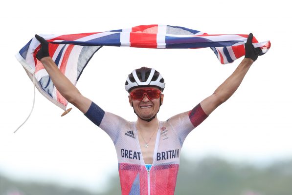 Top Pidcock crosses the line to win Gold at the Tokyo Olympics in 2021, holding a Union Jack above his head and with tears in his eyes