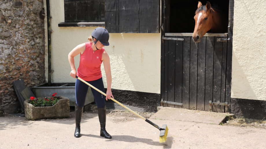Woman sweeping with a yellow outdoor broom outside a stable with a horse looking over the door