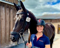 British rider Izzy Palmer pictured wearing a blue T-shirt and riding hat, and holding a dark bay horse