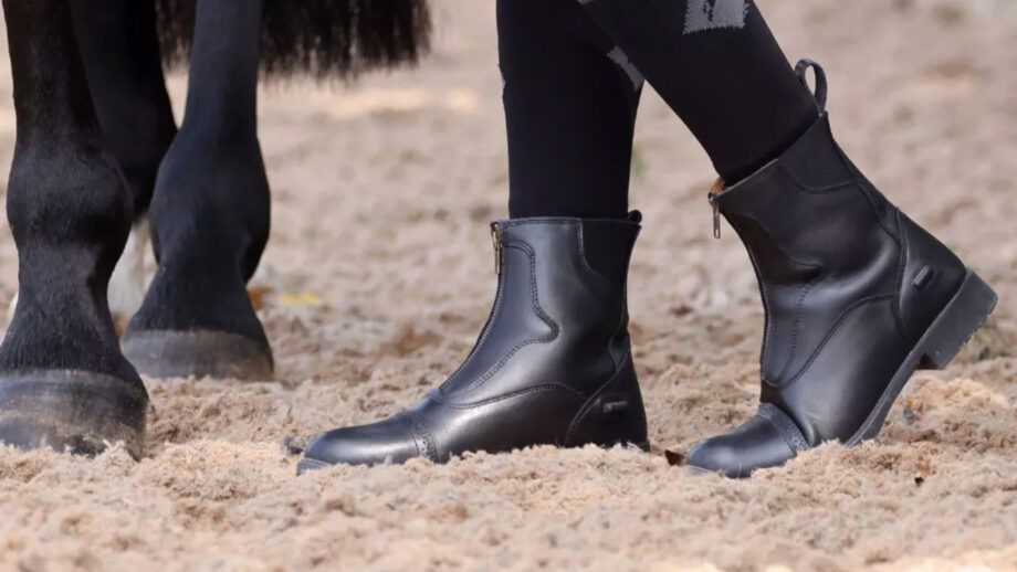 Close up of model wearing short riding boots standing next to a horse in arena