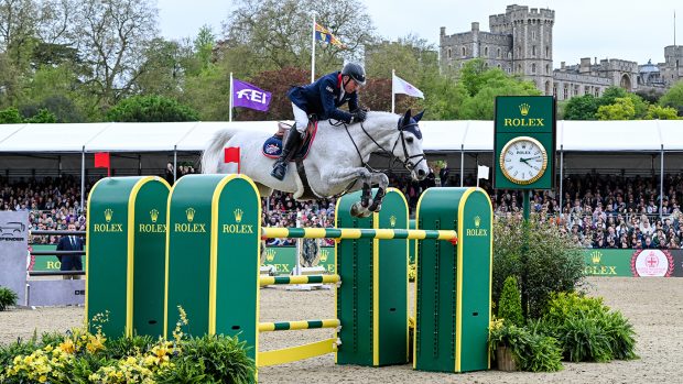 John Whitaker jumping Arqana de Riverland over a large Rolex oxer of green and yellow poles at Royal Windsor Horse Show with Windsor Castle behind the spectator stands