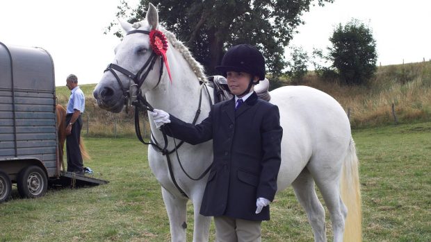 A horsey kid in the noughties dressed an a skullcap and black silk, dark show jacket and cream jodhpurs. He's holding a grey pony, who wears a red rosette.