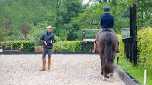 Carl Hester faces the camera while standing in his outdoor school explaining a dressage movement to a pupil who is riding directly away from the camera