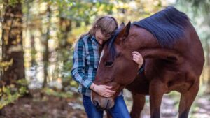 Woman and her bay horse in a forest. The horse is bending his head towards her and she has her arms around him.