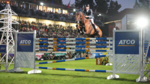 Joe Stockdale jumps a massive sixbar fence towards the camera at Spruce Meadows under the spotlights with the huge stands full of spectators behind.