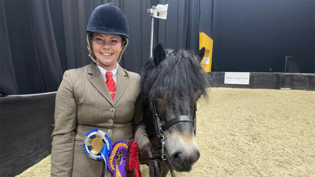 Davina Johnson smiles widely at the camera wearing her winners rosettes on her jacket standing beside her Exmoor pony Stowbrook Red Kite in the backstage area at HOYS 2025.