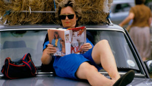 Woman sat on car bonnet at polo match, reading a copy of Jilly Cooper’s Polo