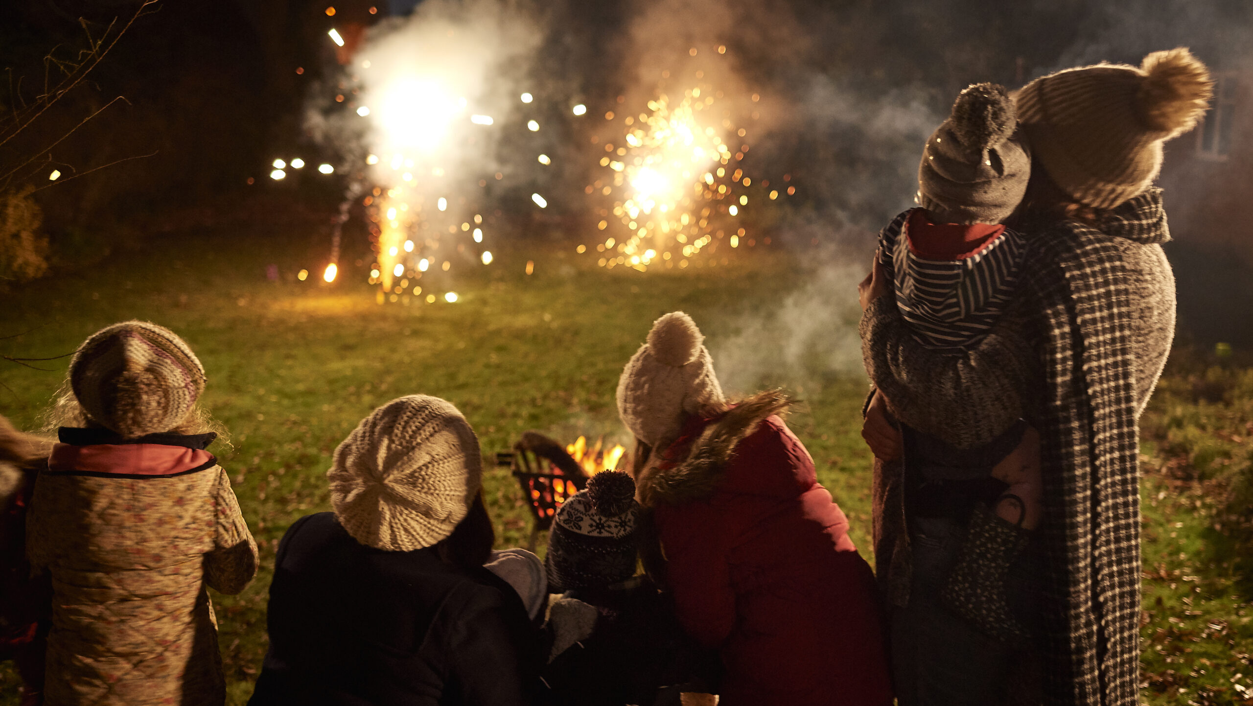 Two young families wrapped up warm, watching home fireworks display in back garden