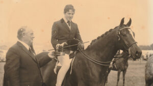 A sepia-toned image of a show pony ridden by Nigel Hollings, who has removed his hat to accept his prize, with the judge standing alongside, who has also removed his hat.