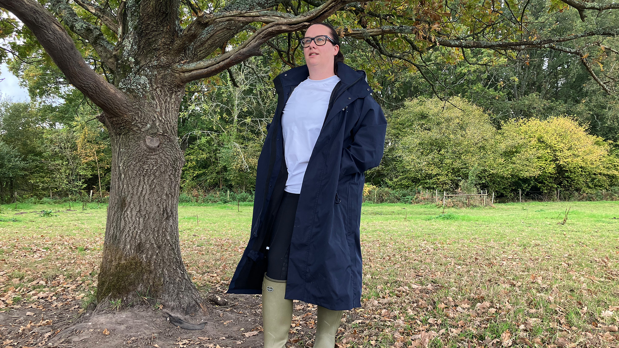 Lady wears Reincoat Lite standing by a tree unzipped, viewed from the front