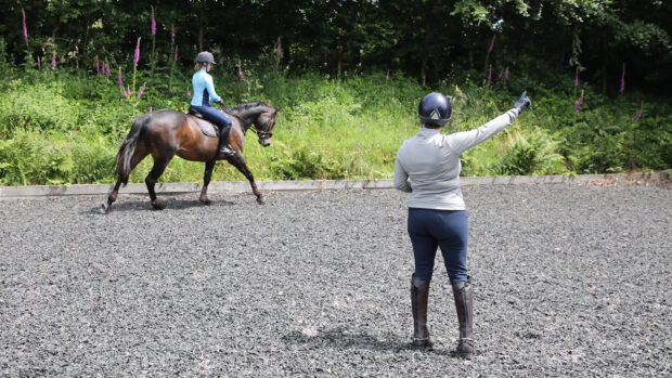 A riding instructor pictured from behind gesturing with her right hand as a adult rider on a bay horse canters on the right rein.