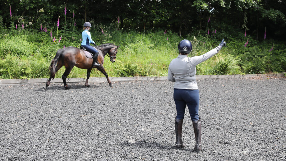 A riding instructor pictured from behind gesturing with her right hand as a adult rider on a bay horse canters on the right rein.