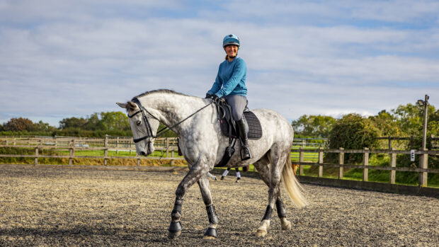 A grey horse walking in an outdoor area with a happy looking adult rider during a riding lesson