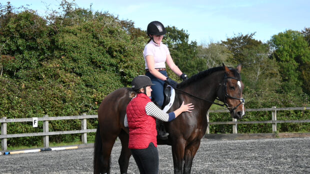 A bay horse at halt with an adult rider on board with a riding insturctor standing alongside with her hand on the horse's shoulder explaining how to ride a horse.