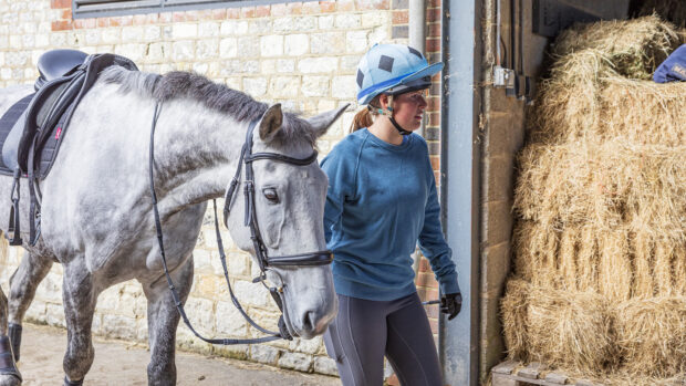 A woman in casual riding clothing with a riding hat and gloves leads a grey horse across a stable yard