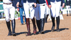 Show jumping horse riders walking the course in a sand arena wearing brown and black leather boots.