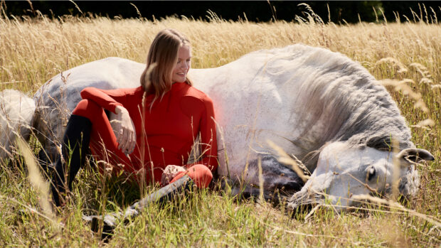 Grey horse lying down in long grass with model sitting down next to it wearing Stella McCartney and LeMieux collection baselayer and breeches in Red