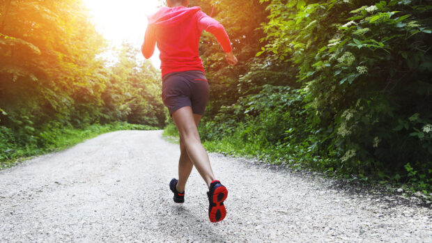 A young woman wearing a red top, dark shorts and red and black trainers running along a country road.