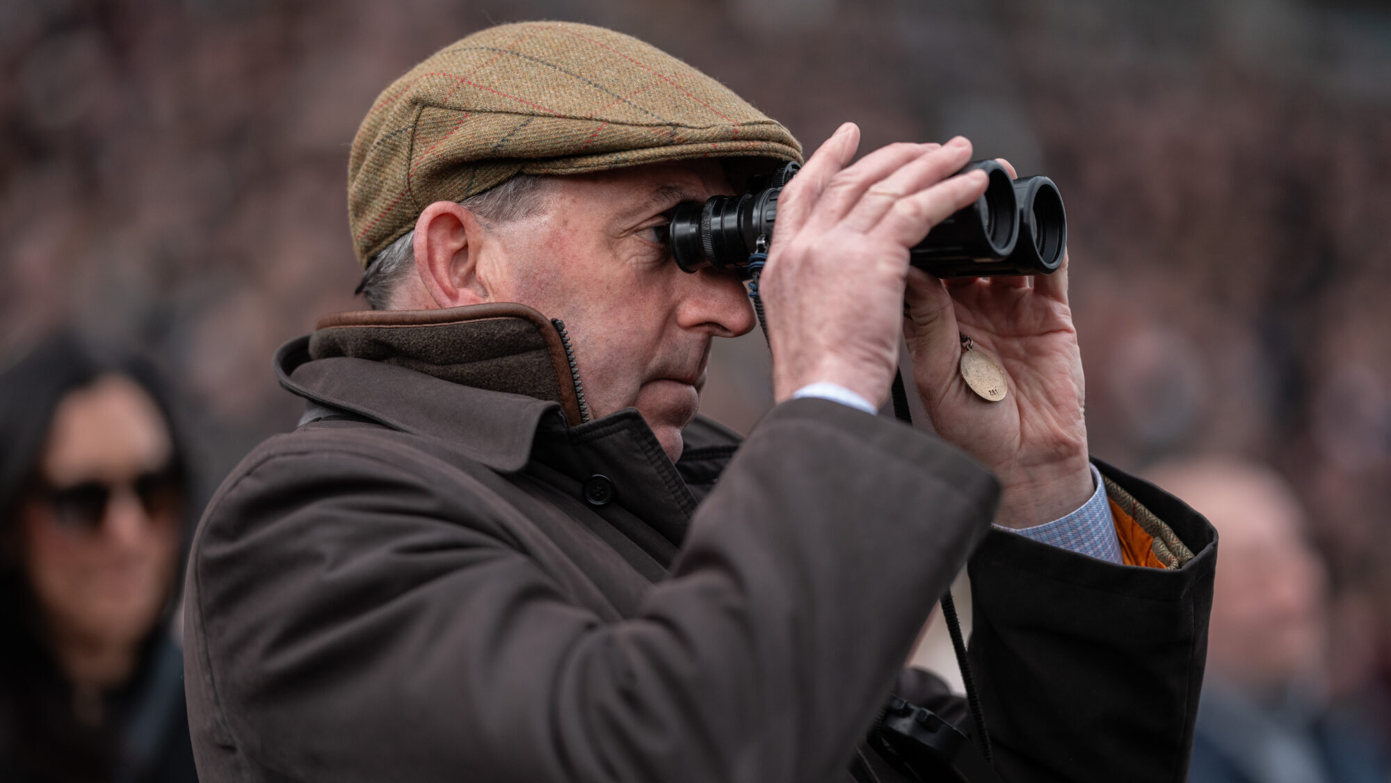Man wearing warm clothing and tweed cap, and looking through binoculars at Cheltenham races