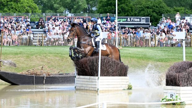 Image shows Ros Canter riding Lordships Graffalo at Badminton Horse Trials in 2025. The pair are jumping a brush arrowhead fence in the water, watched on by crowds
