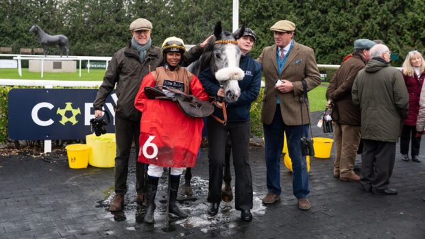 Image shows Aamilah Aswat holding her saddle and red saddle cloth, pictured alongside grey racehorse Guchen and other winning connections at Kempton on Monday (2 February).