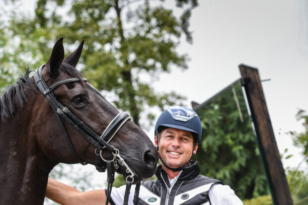 Carl Hester smiles at the camera, stood with En Vogue with arena mirror and trees in the background.