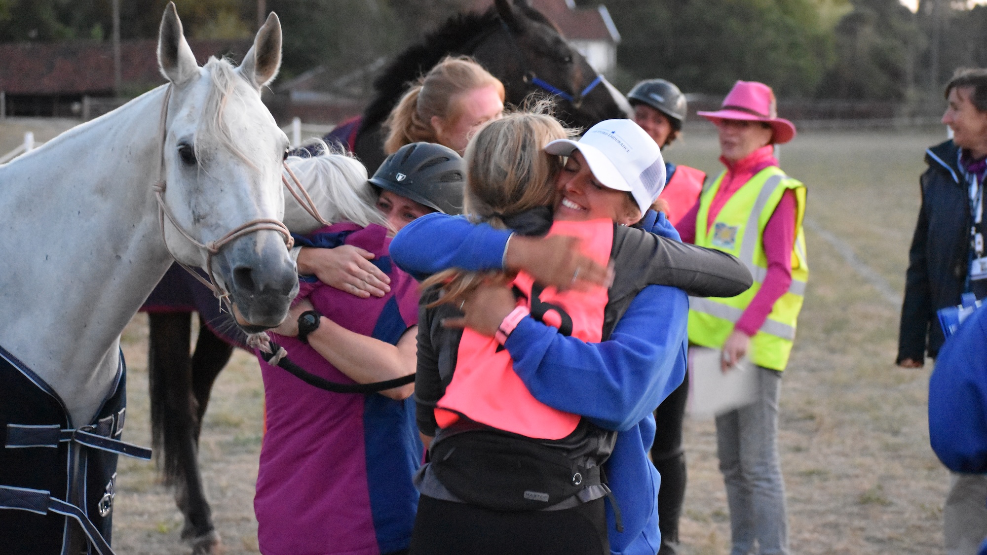 Finishing an endurance race. Riders hugging, grey horse watching on