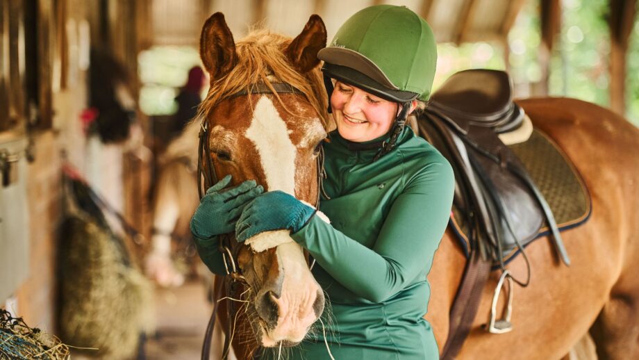 A woman wearing a green baselayer and riding helmet with a green silk cuddles the head of a chestnut horse, who is wearing. saddle and bridle with a sheepskin noseband
