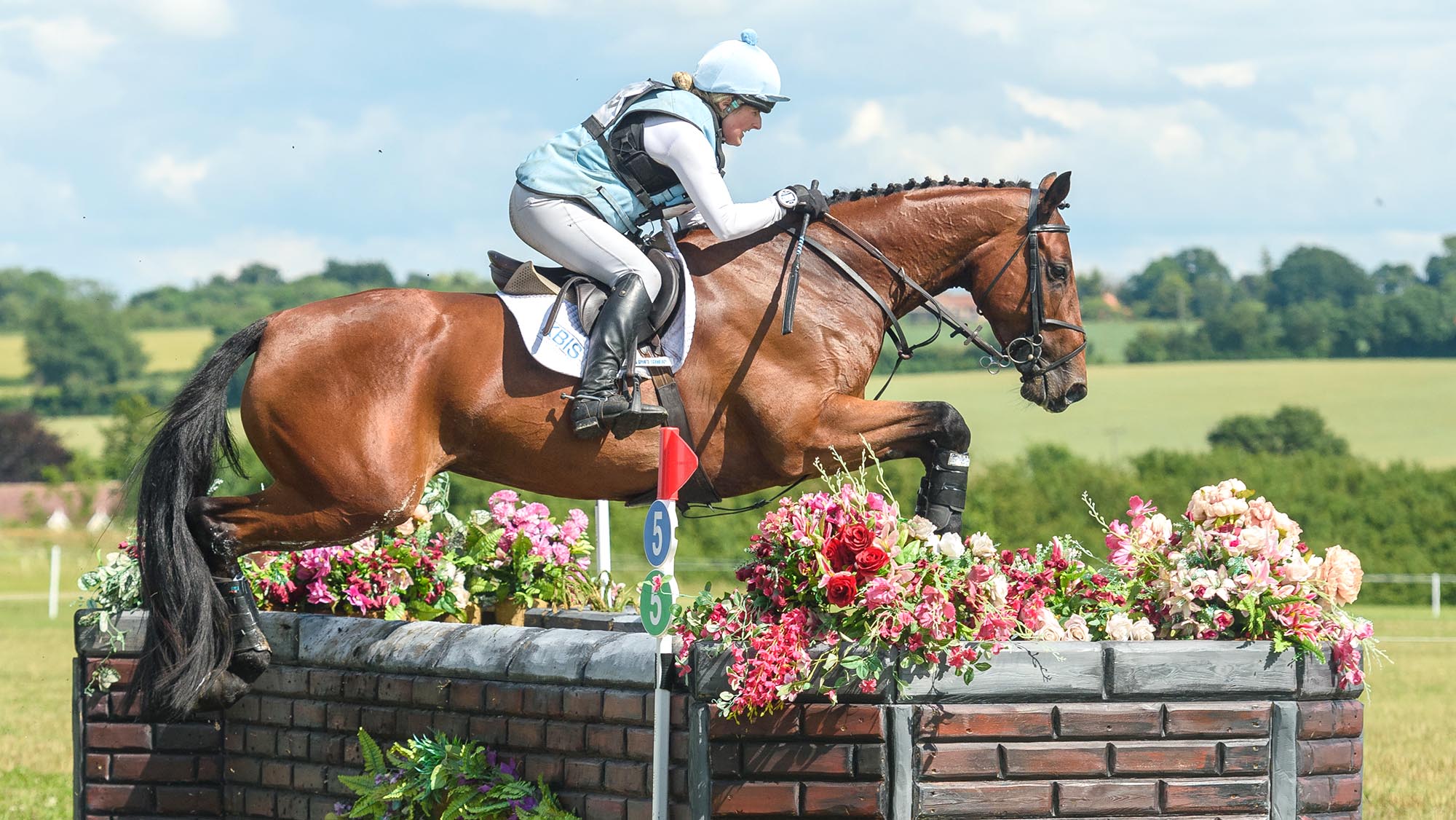 Image shows Sally Butler in her pale blue cross-country colours riding Diamonds Eclipse, who is jumping a corner fence dressed with flowers, on a sunny day at Keysoe in 2021.