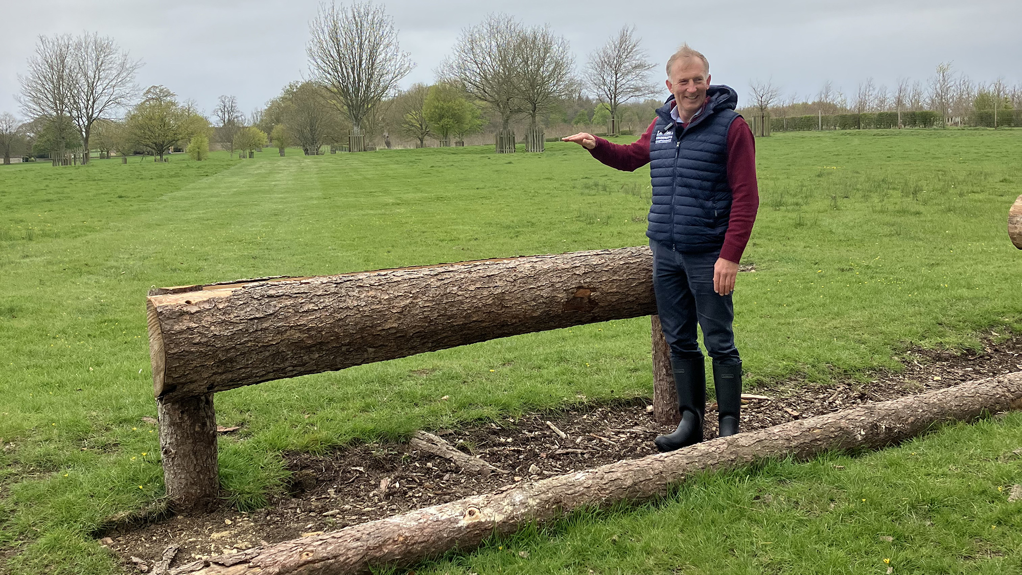 A wooden rail with a scraped out ditch and ground line with the course-designer standing in the ditch holding his raised hand above the rail.