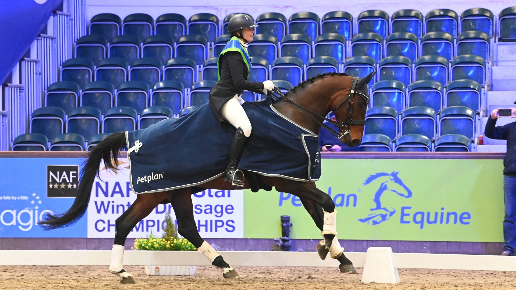Woman wearing a winners sash cantering on a horse who wears a navy winners rug at the winter dressage championships