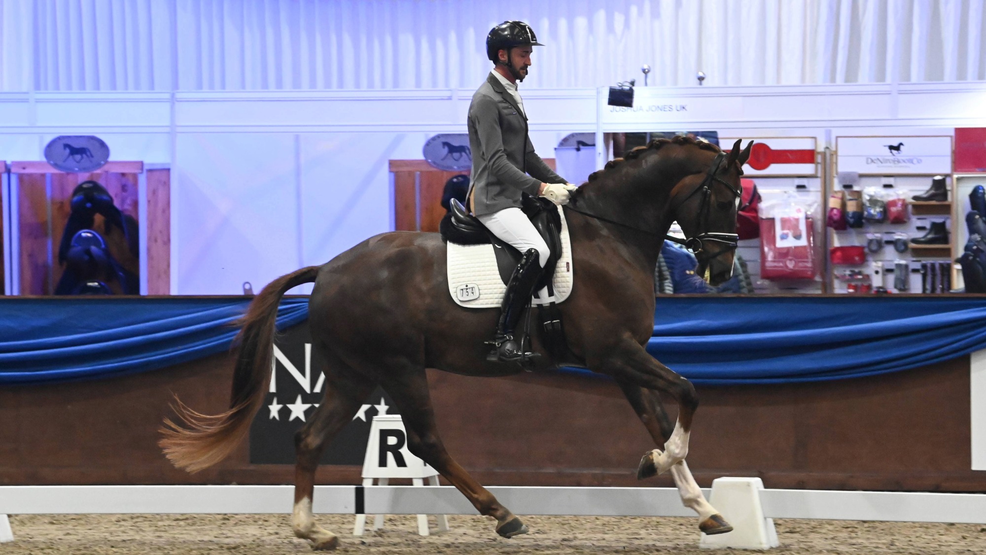 A man, Eli Lovell, cantering on a livery chestnut horse in the indoor arena at the NAF Five Star Winter Dressage Championships