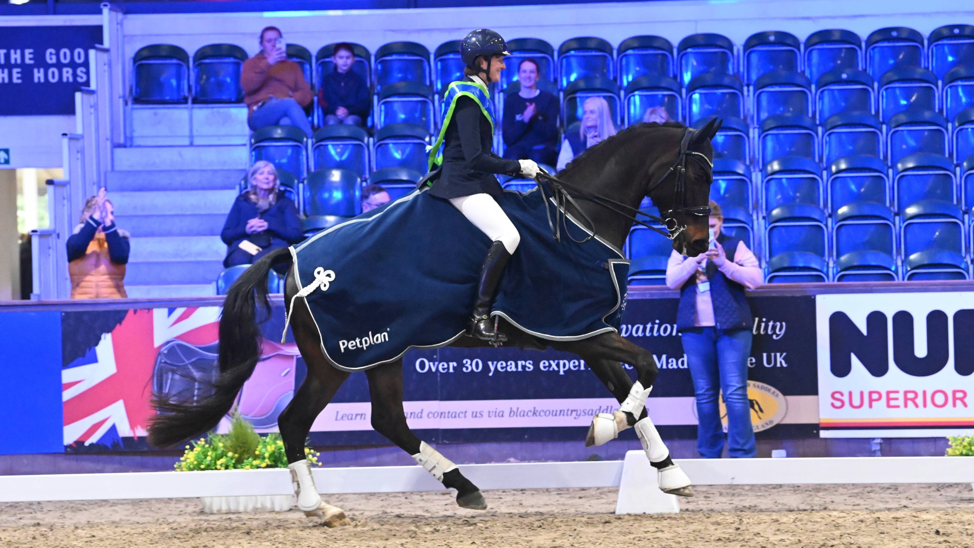 Woman canters a dark bay horse who wears a navy winners rug at the winter dressage championships