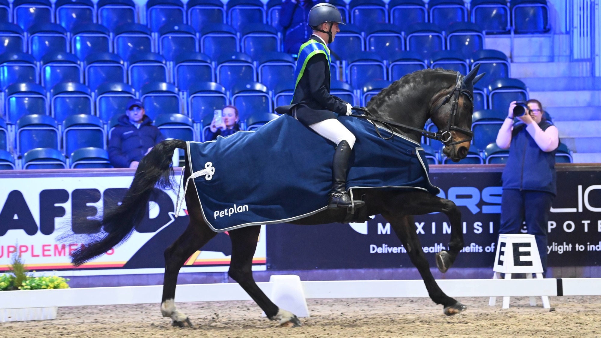 A man rides a black horse at a canter in an indoor arena. The horse wears a blue run and the man a winner's sash. There is blue seating in the stands in the background.