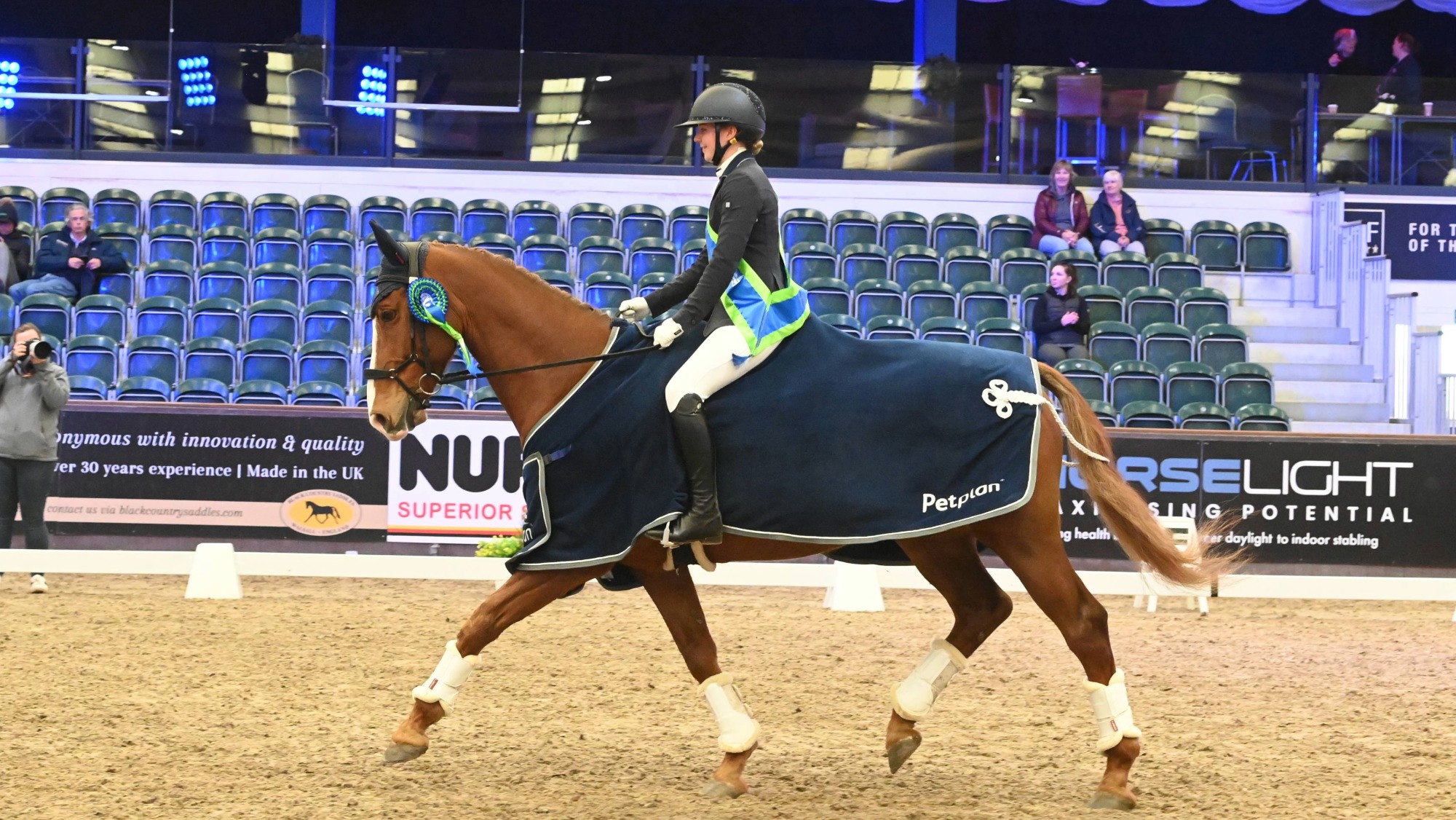 Woman on a chestnut horse who wears a navy rug and rosette. The rider wears a winners sash. They are trotting in an indoor arena.