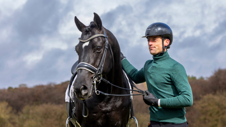 Justin Verboomen wearing a green jumper standing next to his horse Zonik Plus who is tacked up waiting to be ridden