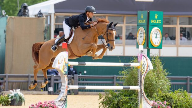 Showjumper Victor Bettendorf wears a black jacket and hat with white breeches on a chestnut horse called Qwando Van De Rispen, clears a large upright jump of black and white poles with a Rolex clock in the background at Royal Windsor Horse Show