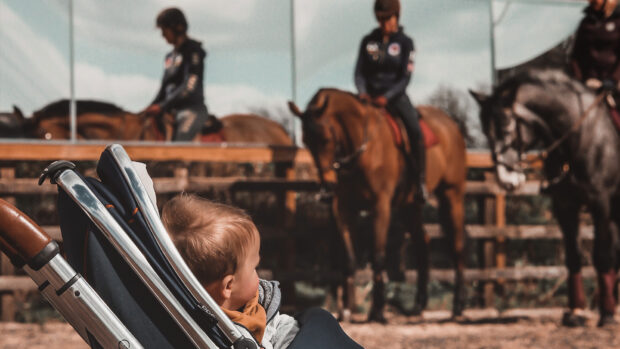 Baby in pram in foreground, with two people riding horses in background after giving birth