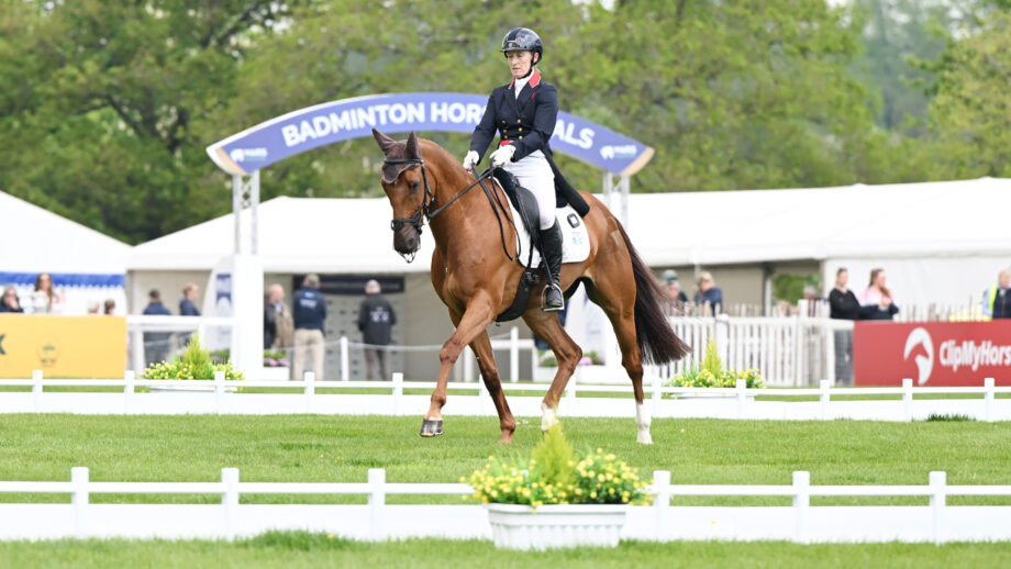 Daisy Berkeley rides a chestnut horse in trot in the grass Badminton Horse Trials dressage arena with the arena entry sign behind her.