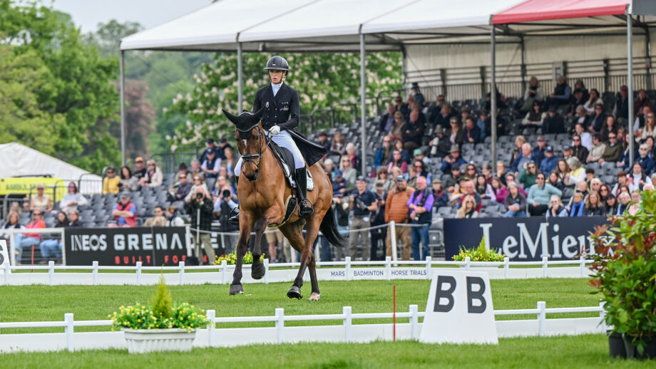 Emily King rides bay horse Valmy Biats in canter towards the camera in the Badminton Horse Trials dressage arena with the packed stands behind.