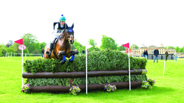 A bay horse clears a large BE100 brush fence on the LeMieux Grassroots course with Badminton House in the background.