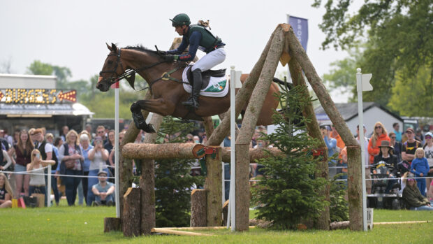 A bay horse jumps a large wooden oxer on the Badminton Horse Trials cross-country course with the crowds in the background