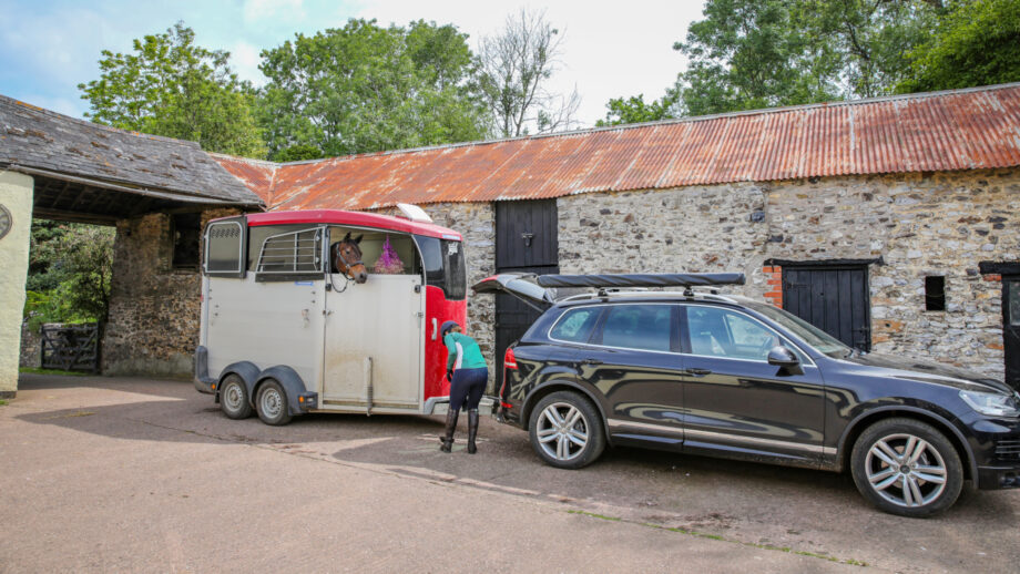 A horsebox on a yard hooked up to a black 4x4. A bay horse can be seen peeroung out of the top window while a woman in a turquoise top packs the car