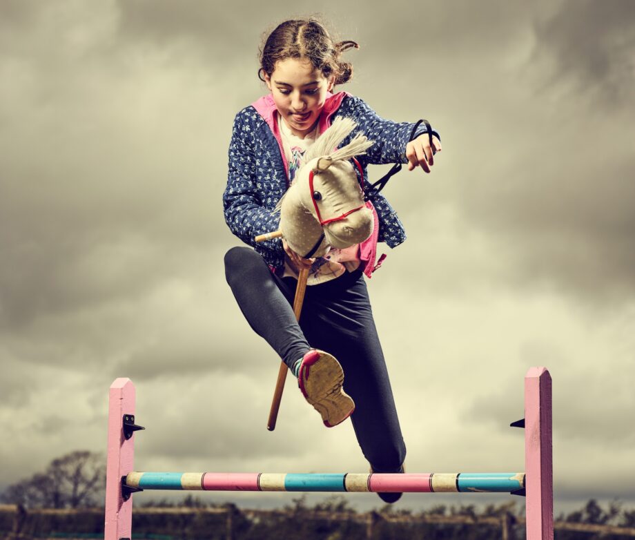 Young girl hobby horsing over a jump, with a cloudy sky above