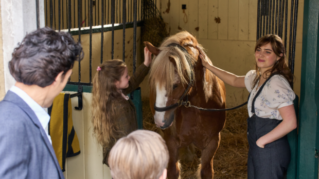 A still from Rivals TV series showing a man and boy looking into a stable where a woman and young girl are caring for a pony.