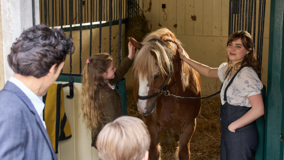 A still from Rivals TV series showing a man and boy looking into a stable where a woman and young girl are caring for a pony.