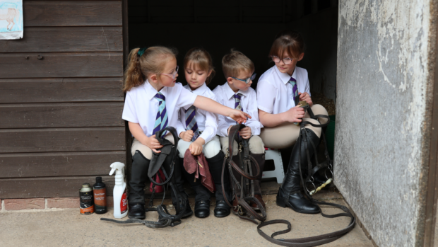 Four children sit in a ack room doorway cleaning tack at Pony Club camp. They wear riding boots, beige jodhpurs, white shirts and blue and purple ties